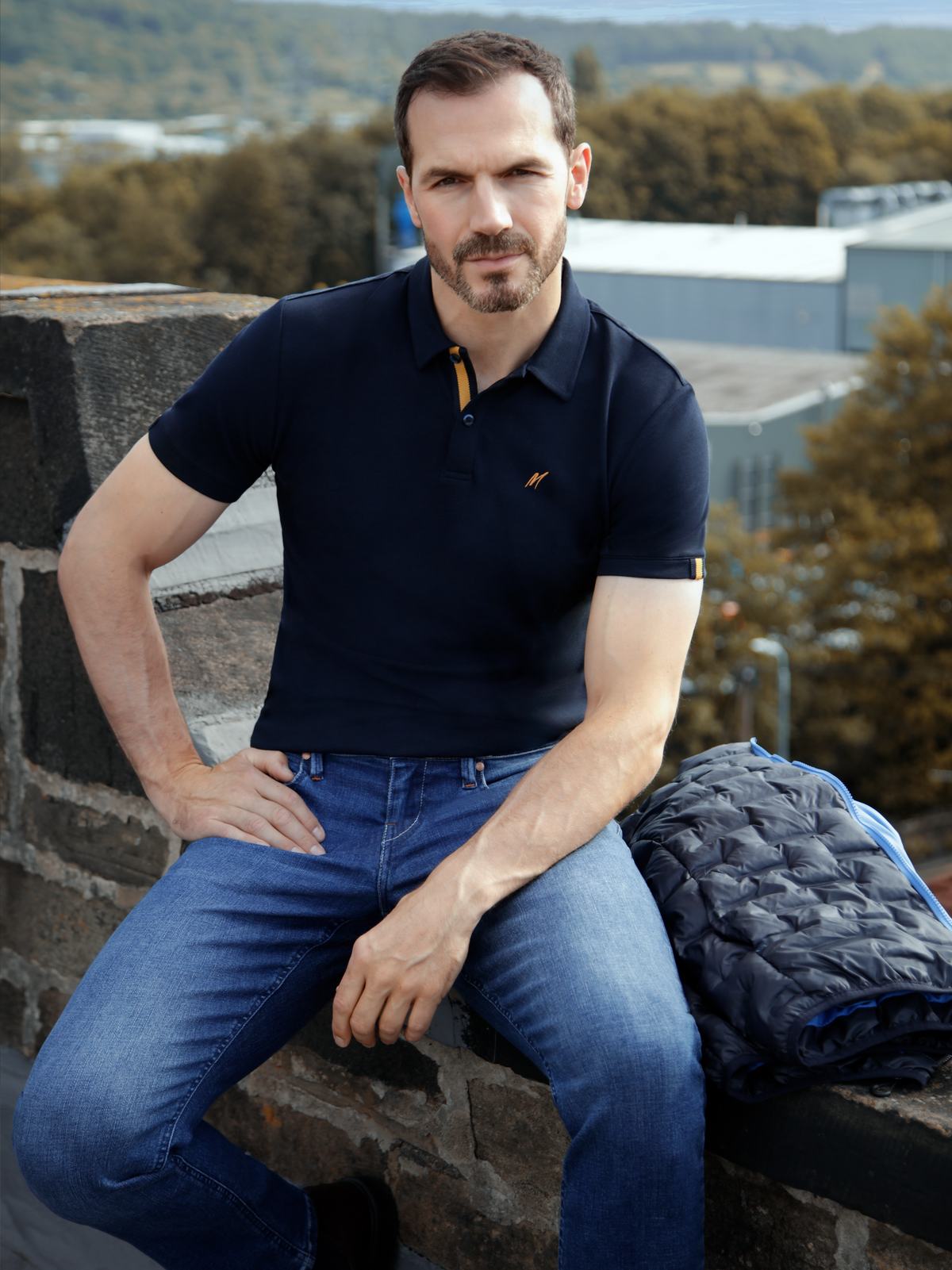 Man wearing Mistral Navy S/S Polo on a rooftop, showcasing a smart casual style with jeans.