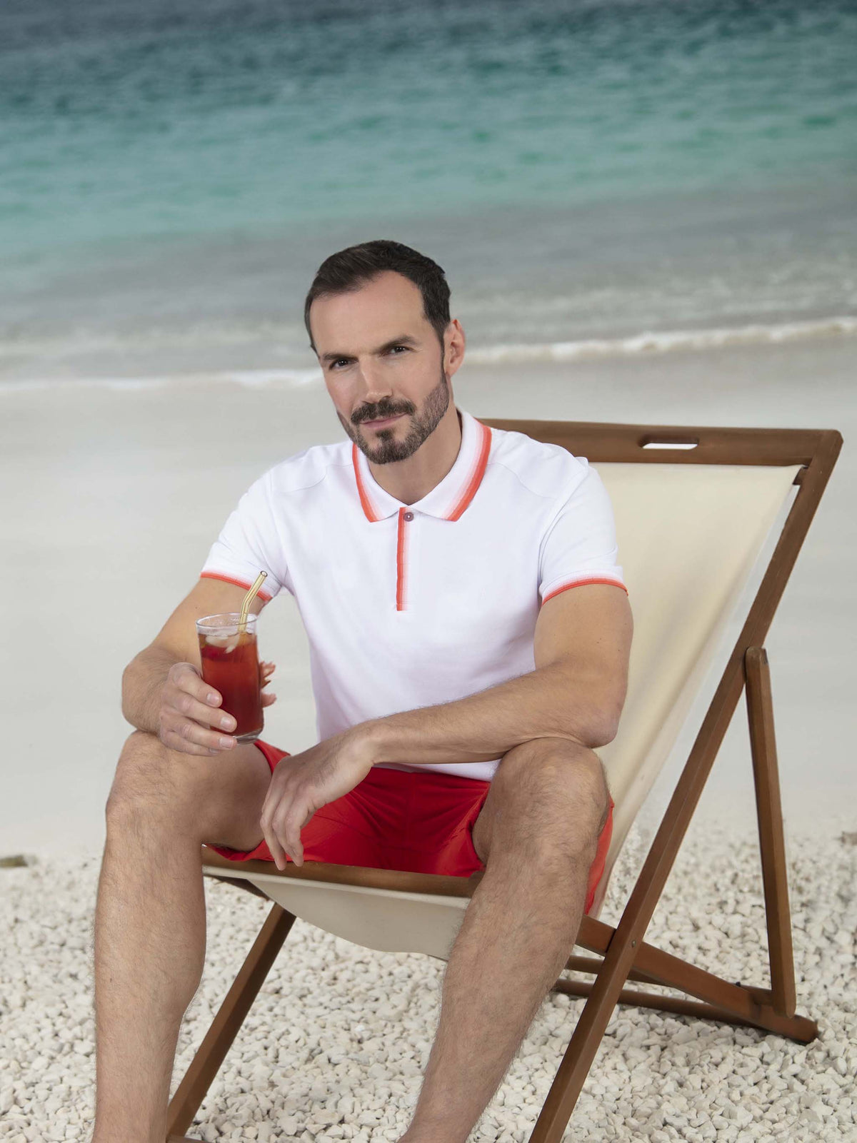 A man sitting on a beach in a white polo and red chino shorts. Menswear UK.